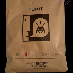 May include: A brown paper bag with the words "ALERT" printed on it. The bag features a black and white graphic of a video game character. The bag also has a tick next to the size "M" and the words "INVADER SPACE STATION 2024" printed on it.