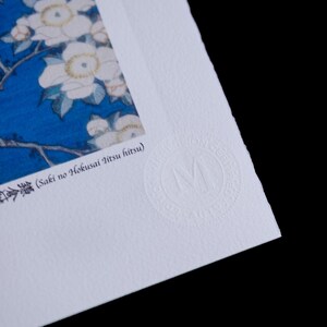 May include: Close-up of a print featuring white flowers with yellow centers against a blue background. The print has Japanese text and a circular embossed logo. The paper has a textured surface and is set against a black background.