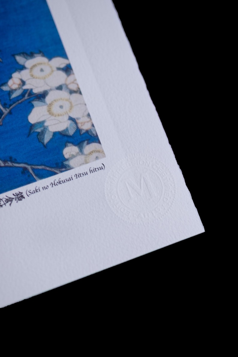 May include: Close-up of a print featuring white flowers with yellow centers against a blue background. The print has Japanese text and a circular embossed logo. The paper has a textured surface and is set against a black background.