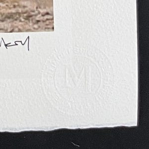 May include: A white textured paper border featuring a handwritten signature in black ink on the left side and a circular embossed stamp with the letter M in the center, positioned below a partial view of a photographic print with earthy tones.