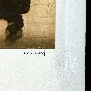 May include: A black and white photograph of a person walking away from the camera. The person is wearing a dark jacket and pants. The background is a cracked wall. The image is signed "Banksy" in the bottom left corner and has a "Made in the UK" stamp in the bottom right corner.