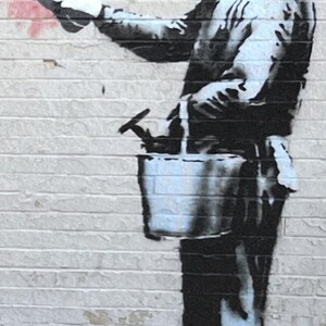Puede incluir: Un mural de estilo estarcido en blanco y negro sobre una pared de ladrillo que representa una figura con gorra y ropa de trabajo. La figura sostiene una esponja y lleva un cubo con herramientas, junto a una marca de pintura roja.