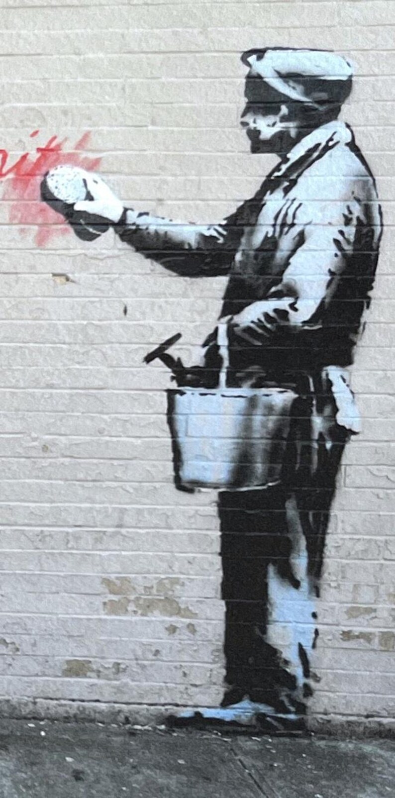 Puede incluir: Un mural de estilo estarcido en blanco y negro sobre una pared de ladrillo que representa una figura con gorra y ropa de trabajo. La figura sostiene una esponja y lleva un cubo con herramientas, junto a una marca de pintura roja.