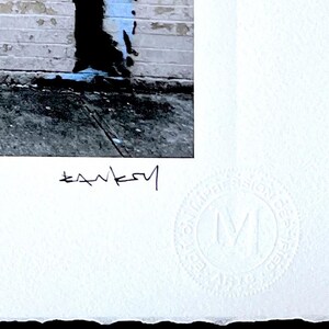 Puede incluir: Una impresi&oacute;n en papel blanco con una imagen fotogr&aacute;fica de una pared de ladrillos con una abertura vertical oscura y detalles en pintura azul. La impresi&oacute;n incluye la firma negra Banksy bajo la imagen y un sello circular en relieve con la letra M.