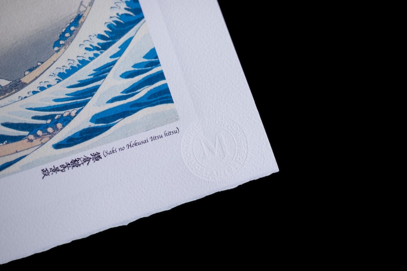 K&ouml;nnte beinhalten: Ein Druck eines japanischen Holzschnittkunstwerks mit einer gro&szlig;en Meereswelle in Blau und Wei&szlig;. Der Druck enth&auml;lt japanischen Text und ein kreisf&ouml;rmiges gepr&auml;gtes Logo. Das Papier hat eine strukturierte Oberfl&auml;che und einen B&uuml;ttenrand.