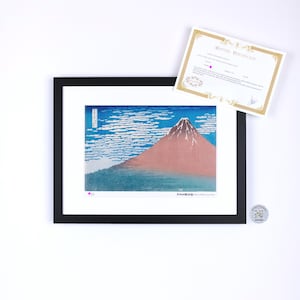 May include: Framed print of a Japanese landscape featuring Mount Fuji with a red peak, blue sky, and white clouds. The print is in a black frame with a white mat. A certificate is included.