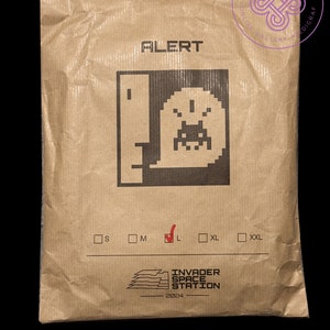May include: A brown paper bag with the text "ALERT" printed on it. The bag features a pixelated image of a spaceship with the text "INVADER SPACE STATION 2024" printed below. The bag has a size chart with checkboxes for S, M, L, XL, and XXL.