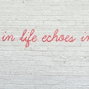 Puede incluir: Una pared de ladrillo blanco con el texto en cursiva roja What we do in life echoes in Eternity. Se ve una parte de una mano con un guante blanco y negro en el lado derecho, sosteniendo un aerosol que est&aacute; aplicando pintura roja.