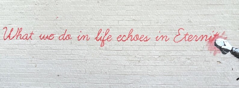 Puede incluir: Una pared de ladrillo blanco con el texto en cursiva roja What we do in life echoes in Eternity. Se ve una parte de una mano con un guante blanco y negro en el lado derecho, sosteniendo un aerosol que est&aacute; aplicando pintura roja.