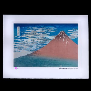 May include: A framed print of a Japanese landscape featuring Mount Fuji. The artwork depicts the mountain in shades of red and brown, set against a blue sky with white cloud formations. The print has Japanese text.