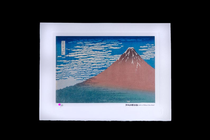 May include: A framed print of a Japanese landscape featuring Mount Fuji. The artwork depicts the mountain in shades of red and brown, set against a blue sky with white cloud formations. The print has Japanese text.
