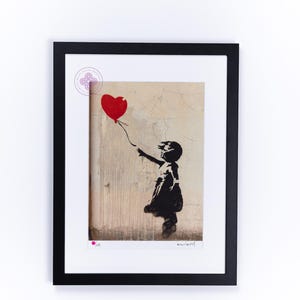 May include: A framed print of a black and white stencil art piece depicting a girl holding a red heart-shaped balloon. The background is a textured, beige wall. The print is signed "Banksy" in the bottom right corner.