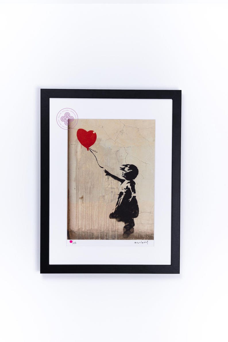 May include: A framed print of a black and white stencil art piece depicting a girl holding a red heart-shaped balloon. The background is a textured, beige wall. The print is signed "Banksy" in the bottom right corner.