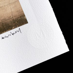 May include: A white paper print with a black signature and a white embossed circle with the letter "M" inside. The circle has the text "Original Limited Edition" around the outside.