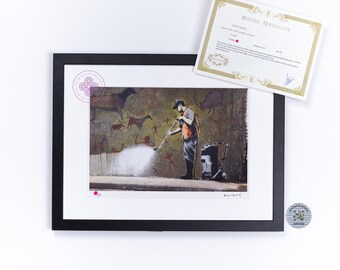 May include: A framed art print featuring a person in an orange vest using a pressure washer to clean a wall covered in prehistoric-style cave paintings. The print includes a white mat, a black frame, and a separate document titled Editing Certificate.