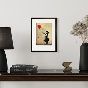 May include: A framed print of a black and white stencil art piece depicting a girl holding a red heart-shaped balloon. The background is a faded, textured beige.