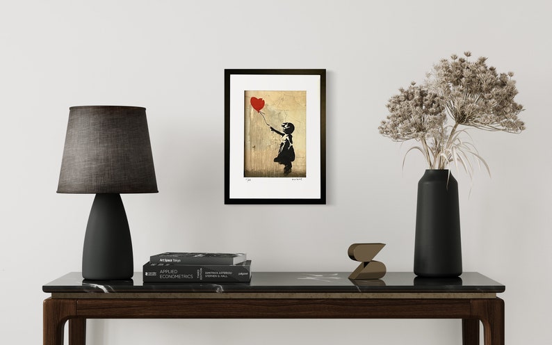 May include: A framed print of a black and white stencil art piece depicting a girl holding a red heart-shaped balloon. The background is a faded, textured beige.