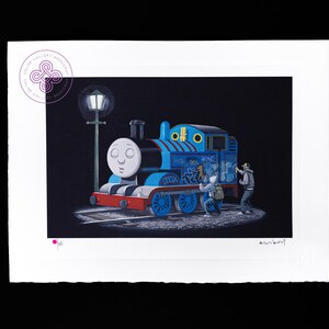 May include: A rectangular art print on white paper featuring a blue steam engine train with a white face, red bumper, and blue wheels. Two figures are spray painting graffiti on the side of the train under a street lamp against a black background.