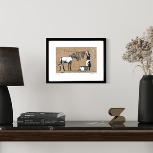 May include: A framed art print featuring a black and white illustration of a zebra standing near a person hanging laundry on a clothesline against a tan brick wall. The print is displayed in a simple black frame on a white wall.