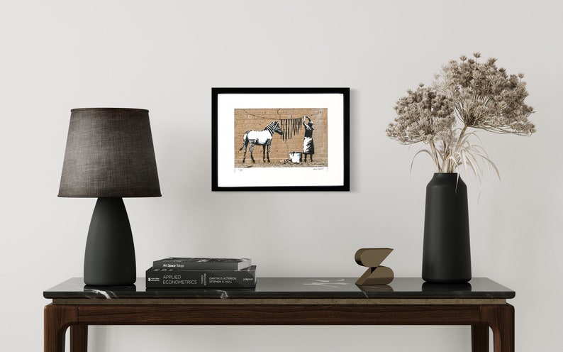 May include: A framed art print featuring a black and white illustration of a zebra standing near a person hanging laundry on a clothesline against a tan brick wall. The print is displayed in a simple black frame on a white wall.