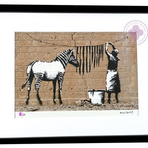 May include: A framed print featuring a black and white stencil-style artwork of a zebra standing next to a person hanging black strips on a clothesline against a tan brick wall. A bucket sits on the ground. The print includes a signature and numbering.