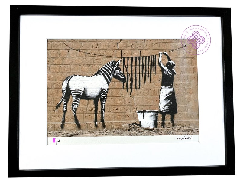 May include: A framed print featuring a black and white stencil-style artwork of a zebra standing next to a person hanging black strips on a clothesline against a tan brick wall. A bucket sits on the ground. The print includes a signature and numbering.