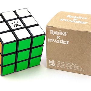 May include: A black, white, orange, and green Rubik's Cube with a Space Invaders graphic on one side. The cube is next to a brown box with the text "Rubik's x Invader" and "Limited Edition for the exhibition Invader Rubikcubiste at MIMA - Brussels 2022".