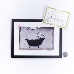Peut inclure: Une estampe encadrée en noir et blanc d'une œuvre de street art représentant une figure dans une baignoire tenant une pagaie. L'estampe est signée "Banksy" en bas à droite.