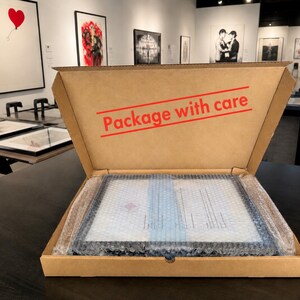 May include: A brown cardboard shipping box with the text "Package with care" printed in red on the inside lid. The box contains a rectangular item wrapped in clear bubble wrap, positioned flat within the base of the container.