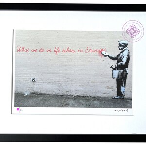 Puede incluir: Una impresi&oacute;n enmarcada que presenta una imagen en blanco y negro estilo plantilla de una persona sosteniendo un cubo y pintando el texto rojo "What we do in life echoes in Eternity" en una pared de ladrillo. Incluye firma y un cuadrado rosa.