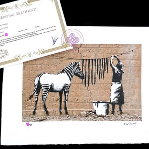 May include: A Banksy art print titled Zebra featuring a black and white zebra standing next to a figure hanging black stripes on a clothesline against a brick wall, accompanied by an Editing Certificate with the text Author: Banksy, Name of the work: Zebra.