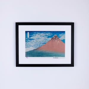 May include: Framed print of a Japanese landscape featuring Mount Fuji. The artwork depicts the mountain with a snow-capped peak, set against a blue sky with white cloud formations. The print is in a black frame.