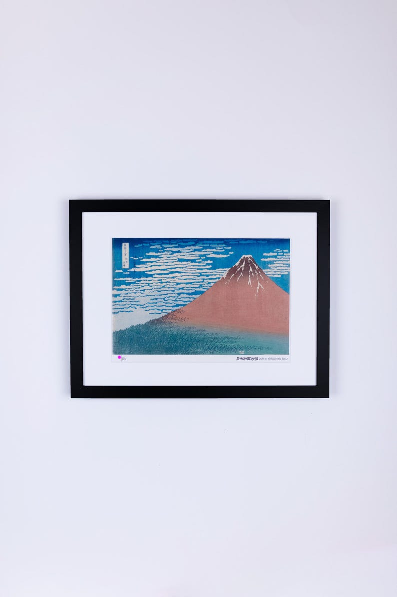 May include: Framed print of a Japanese landscape featuring Mount Fuji. The artwork depicts the mountain with a snow-capped peak, set against a blue sky with white cloud formations. The print is in a black frame.