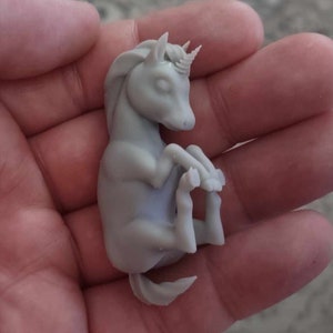 May include: A small, gray, 3D printed unicorn figurine with a curled tail and a horn. The unicorn is lying on its side with its head resting on its front legs.