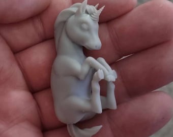 Puede incluir: Una pequeña figura de unicornio de color gris, impresa en 3D, con una cola rizada y un cuerno. El unicornio está acostado de lado con la cabeza apoyada en sus patas delanteras.
