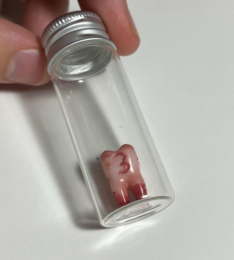 K&ouml;nnte beinhalten: Ein klares Glasfl&auml;schchen mit silbernem Deckel, das einen Zahn enth&auml;lt. Der Zahn ist rosa mit roten Akzenten und die Zahl "3" ist darauf geschrieben. Das Fl&auml;schchen wird von einer Person gehalten.