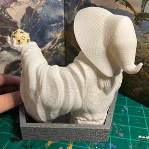 以下が含まれることがあります： 白いD20のサイコロを持ち、9、11、13の数字が見える、白い3Dプリントされた魔法使いの置物。魔法使いは大きな尖った帽子と長いローブを着ています。置物は灰色の石の台座の上に座っています。