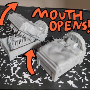 Puede incluir: Dos camas grises impresas en 3D, una con una tapa con bisagras que se abre para revelar una boca de monstruo llena de dientes. El texto "MOUTH OPENS!" está escrito en rojo y negro sobre las camas.