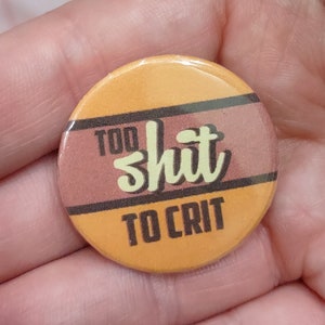May include: A round button with the text "TOO SHIT TO CRIT" in a playful font. The button has a yellow and brown color scheme with a glossy finish. The text is in a light green color.