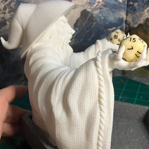 Op de afbeelding: Witte plastic figuur van een tovenaar met een puntige hoed die twee dobbelstenen vasthoudt. De dobbelstenen zijn geel met zwarte cijfers. De figuur zit op een grijze basis.