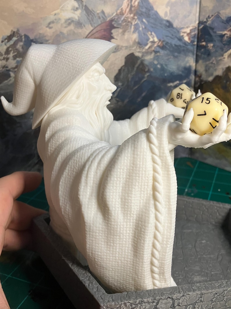 Op de afbeelding: Witte plastic figuur van een tovenaar met een puntige hoed die twee dobbelstenen vasthoudt. De dobbelstenen zijn geel met zwarte cijfers. De figuur zit op een grijze basis.