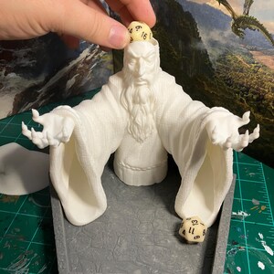 Op de afbeelding: Witte 3D-geprinte dobbelsteen toren in de vorm van een tovenaar met een baard en uitgestrekte armen. De toren heeft een grijze basis met een getextureerd oppervlak. Twee dobbelstenen zijn zichtbaar, &eacute;&eacute;n bovenop het hoofd van de tovenaar en &eacute;&eacute;n in de basis van de toren.