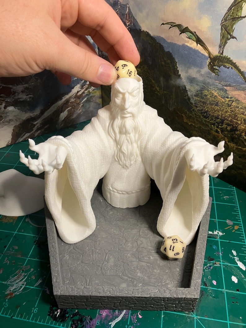 Op de afbeelding: Witte 3D-geprinte dobbelsteen toren in de vorm van een tovenaar met een baard en uitgestrekte armen. De toren heeft een grijze basis met een getextureerd oppervlak. Twee dobbelstenen zijn zichtbaar, &eacute;&eacute;n bovenop het hoofd van de tovenaar en &eacute;&eacute;n in de basis van de toren.