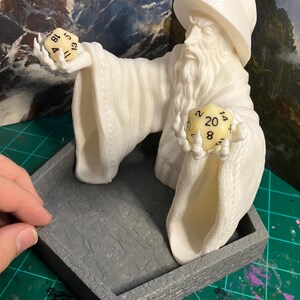 以下が含まれることがあります： 白い3Dプリントされた魔法使いの置物で、2つのサイコロを持っています。魔法使いは白いローブととがった帽子をかぶっています。サイコロは20面サイコロと12面サイコロです。置物は灰色の六角形の台座の上に置かれています。