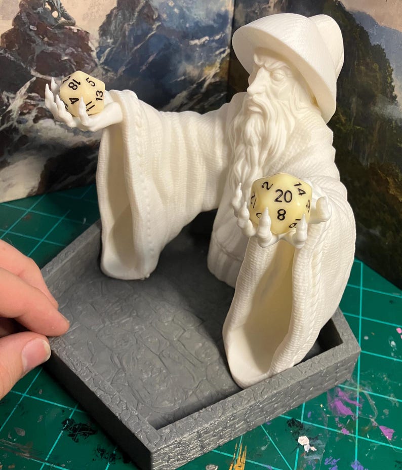 以下が含まれることがあります： 白い3Dプリントされた魔法使いの置物で、2つのサイコロを持っています。魔法使いは白いローブととがった帽子をかぶっています。サイコロは20面サイコロと12面サイコロです。置物は灰色の六角形の台座の上に置かれています。