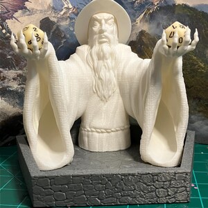 以下が含まれることがあります： 白いプラスチック製の魔法使いの置物。長い白いひげを生やし、白いローブと尖った帽子をかぶっています。両手に黄色いサイコロを2つずつ持っています。置物は灰色の石の台座の上に置かれています。