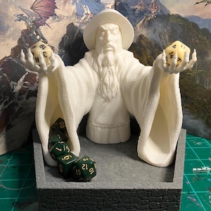 May include: White plastic figurine of a wizard holding two dice, one yellow and one green, with a grey stone base and a green felt insert. The wizard is wearing a robe and has a long white beard. The dice are numbered 1-6.