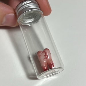 May include: A clear glass vial with a silver cap containing a tooth. The tooth is pink with red accents and the number "3" is written on it. The vial is held by a person.