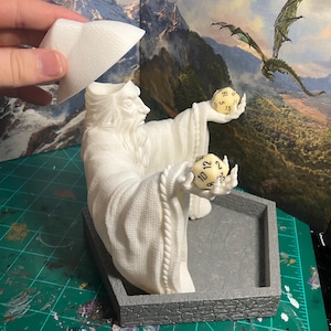 Op de afbeelding: Witte plastic figuur van een tovenaar die twee dobbelstenen vasthoudt. De tovenaar draagt een robe en een hoed. De dobbelstenen zijn genummerd van 1 tot 12 en van 1 tot 20. De figuur staat op een grijze plastic basis.