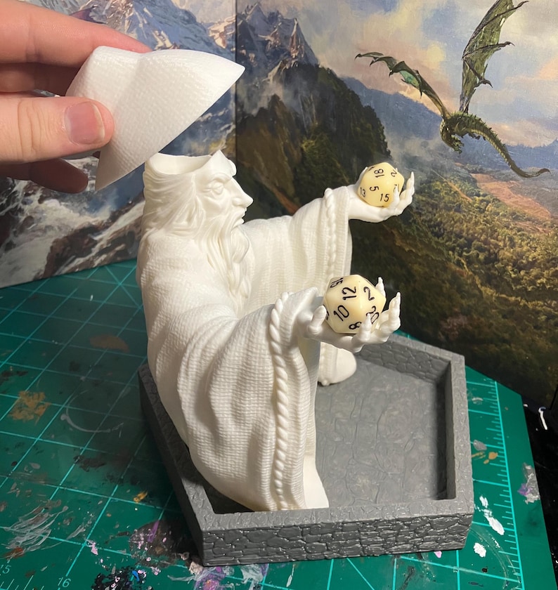 Op de afbeelding: Witte plastic figuur van een tovenaar die twee dobbelstenen vasthoudt. De tovenaar draagt een robe en een hoed. De dobbelstenen zijn genummerd van 1 tot 12 en van 1 tot 20. De figuur staat op een grijze plastic basis.